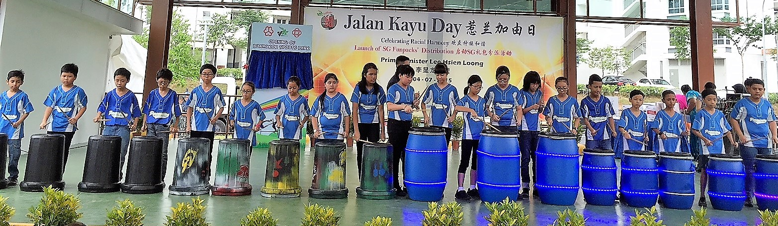 Children in blue uniforms drum on barrels on a stage at Jalan Kayu Day event celebrating Racial Harmony.